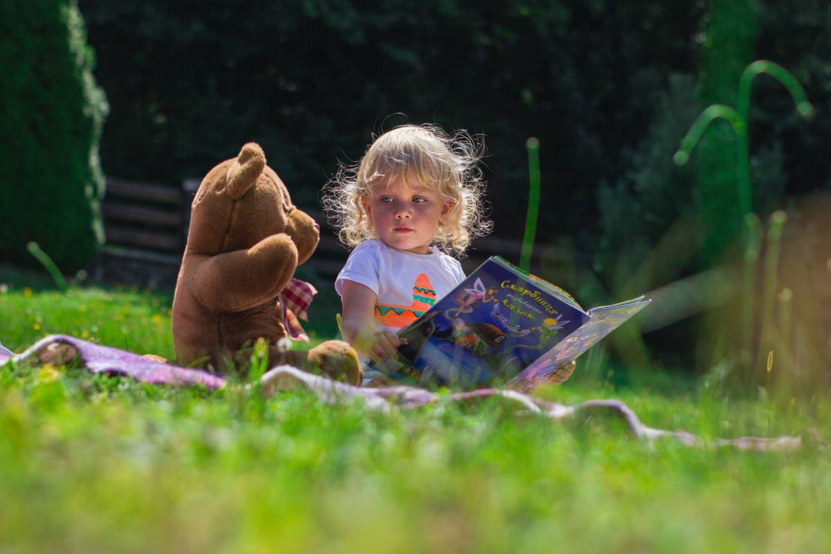 A toddler looking the book with animals and looking to his or her toy bear.